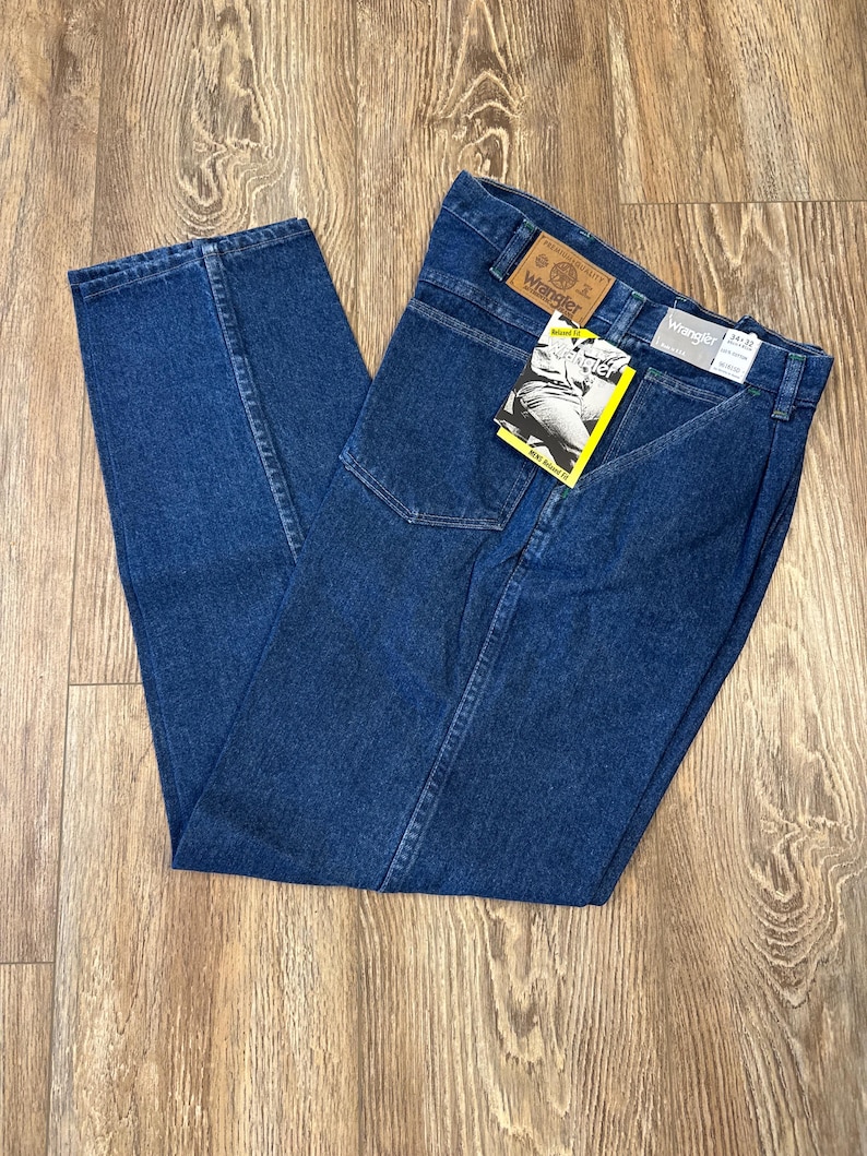 May include: Blue denim Wrangler jeans with a straight leg cut. The jeans have a classic five-pocket design, a brown leather patch, and a yellow tag. The jeans are folded on a wooden surface.