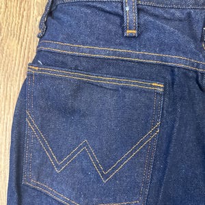 May include: Close-up of dark blue denim jeans. The back pocket features a distinctive 'W' shaped stitching in a contrasting yellow-orange thread. The jeans have a classic five-pocket design with visible stitching and a sturdy waistband.
