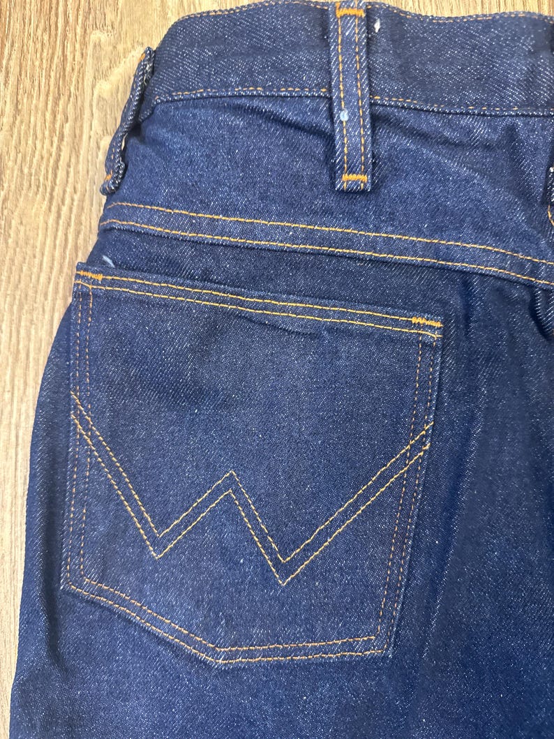 May include: Close-up of dark blue denim jeans. The back pocket features a distinctive 'W' shaped stitching in a contrasting yellow-orange thread. The jeans have a classic five-pocket design with visible stitching and a sturdy waistband.
