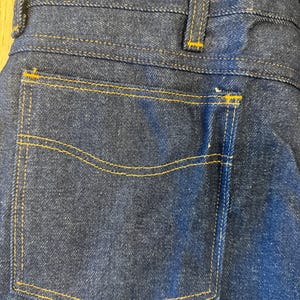 May include: Close-up of the back of dark blue denim jeans. The jeans feature a back pocket with yellow stitching, and the seams are also highlighted with yellow thread. The waistband and belt loops are visible at the top.
