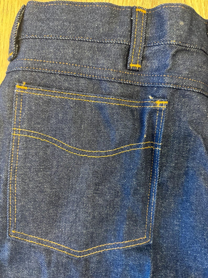 May include: Close-up of the back of dark blue denim jeans. The jeans feature a back pocket with yellow stitching, and the seams are also highlighted with yellow thread. The waistband and belt loops are visible at the top.