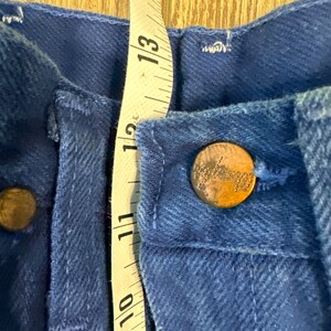 May include: Close-up of a pair of blue denim jeans with a metal button and a measuring tape. The tape measures 10 to 14 inches.