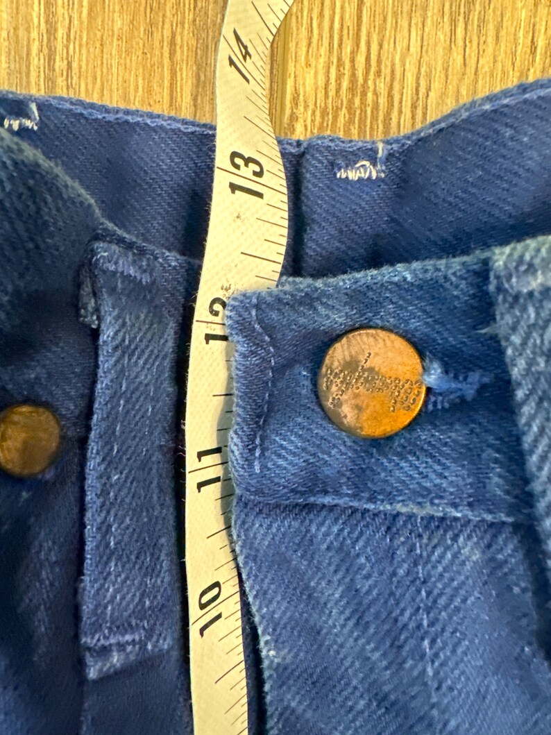 May include: Close-up of a pair of blue denim jeans with a metal button and a measuring tape. The tape measures 10 to 14 inches.