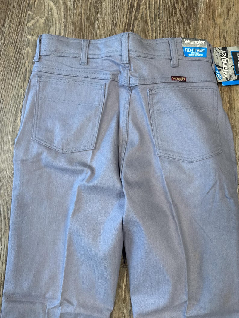 May include: Light blue Wrangler Flex Fit waist jeans. The back view shows the pockets, belt loops, and the Wrangler logo. The tag reads "FLEX-FIT WAIST" for added comfort. The jeans are made of a durable fabric.