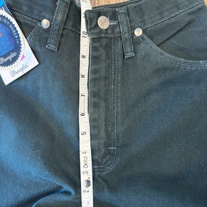 May include: A pair of dark green Wrangler jeans with a measuring tape showing the waist measurement is 10 inches. The jeans have a classic five-pocket style and a straight leg.