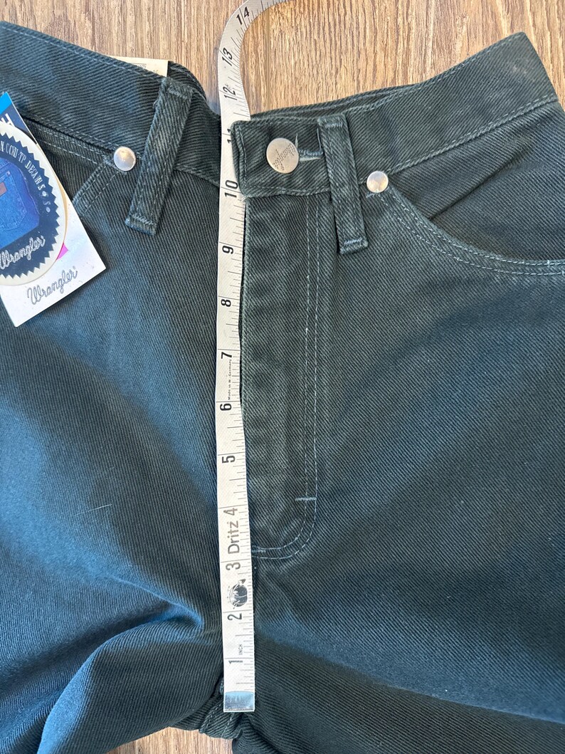 May include: A pair of dark green Wrangler jeans with a measuring tape showing the waist measurement is 10 inches. The jeans have a classic five-pocket style and a straight leg.