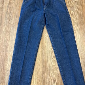 May include: A pair of blue denim jeans with a zipper fly and pleats at the waist. The jeans are new with tags and have a classic, straight-leg fit. The denim is a medium wash, and the jeans are laid flat on a wooden surface.