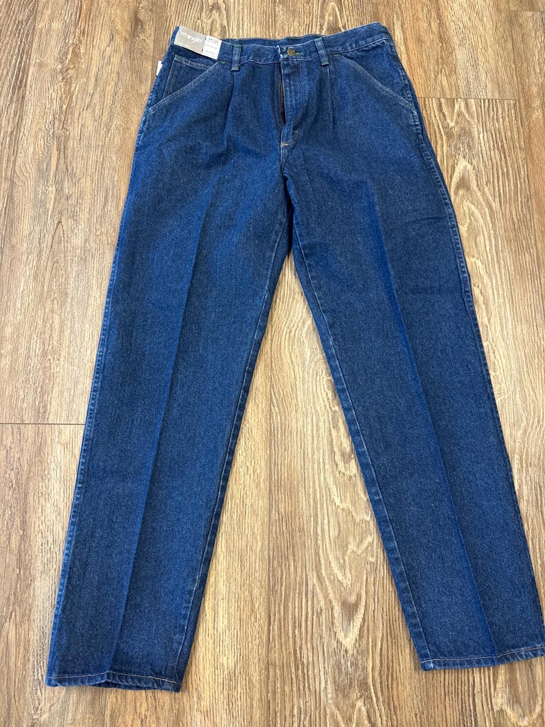May include: A pair of blue denim jeans with a zipper fly and pleats at the waist. The jeans are new with tags and have a classic, straight-leg fit. The denim is a medium wash, and the jeans are laid flat on a wooden surface.