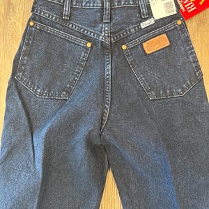 May include: Dark wash denim Wrangler jeans. The jeans have a classic five-pocket design with a brown leather patch and gold-colored rivets. A price tag is attached. The jeans are size 7/8.