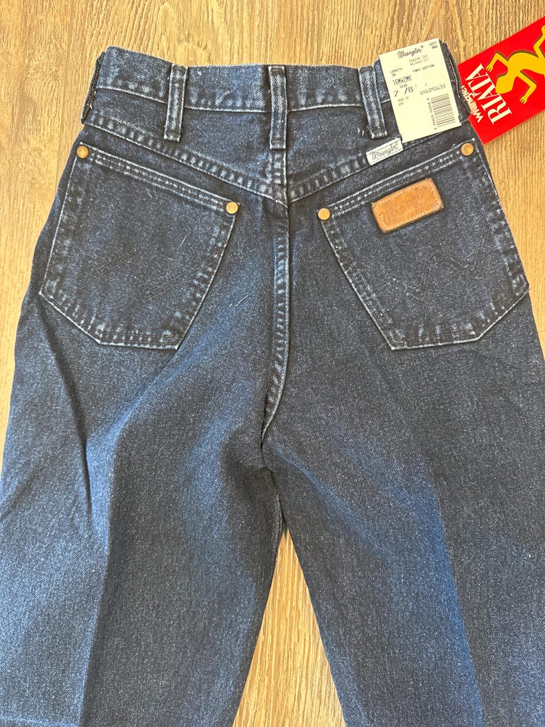 May include: Dark wash denim Wrangler jeans. The jeans have a classic five-pocket design with a brown leather patch and gold-colored rivets. A price tag is attached. The jeans are size 7/8.