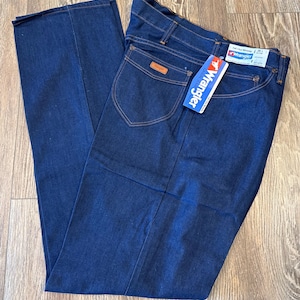 May include: A pair of dark blue denim jeans with a brown leather patch on the back pocket. The jeans have a Wrangler tag attached to the waistband.