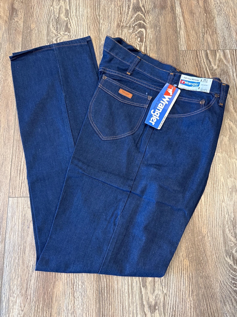 May include: A pair of dark blue denim jeans with a brown leather patch on the back pocket. The jeans have a Wrangler tag attached to the waistband.