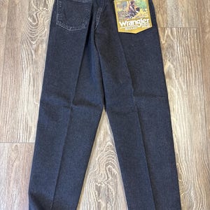 Może przedstawiać: Ciemne dżinsy z praniem z luźnym krojem. Dżinsy mają klasyczny pięciokieszeniowy design, metkę Wrangler na tylnej kieszeni i zawieszkę. Dżinsy są prezentowane na drewnianej powierzchni.