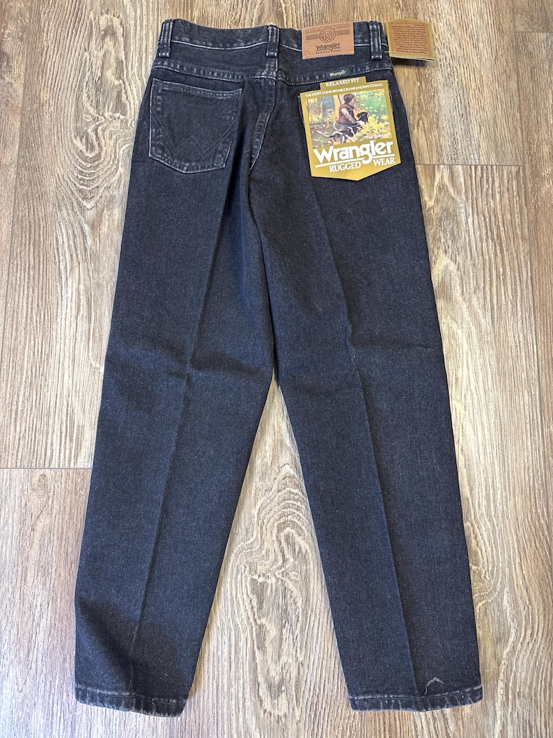 Może przedstawiać: Ciemne dżinsy z praniem z luźnym krojem. Dżinsy mają klasyczny pięciokieszeniowy design, metkę Wrangler na tylnej kieszeni i zawieszkę. Dżinsy są prezentowane na drewnianej powierzchni.