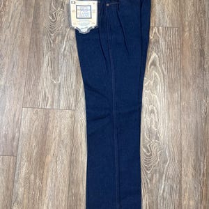 May include: Dark blue Wrangler 915 Classic Jeans, folded and displayed on a wooden surface. The jeans have a classic five-pocket design, a leather patch, and a tag that reads "915 Classic Jeans". The size tag reads 34x34.