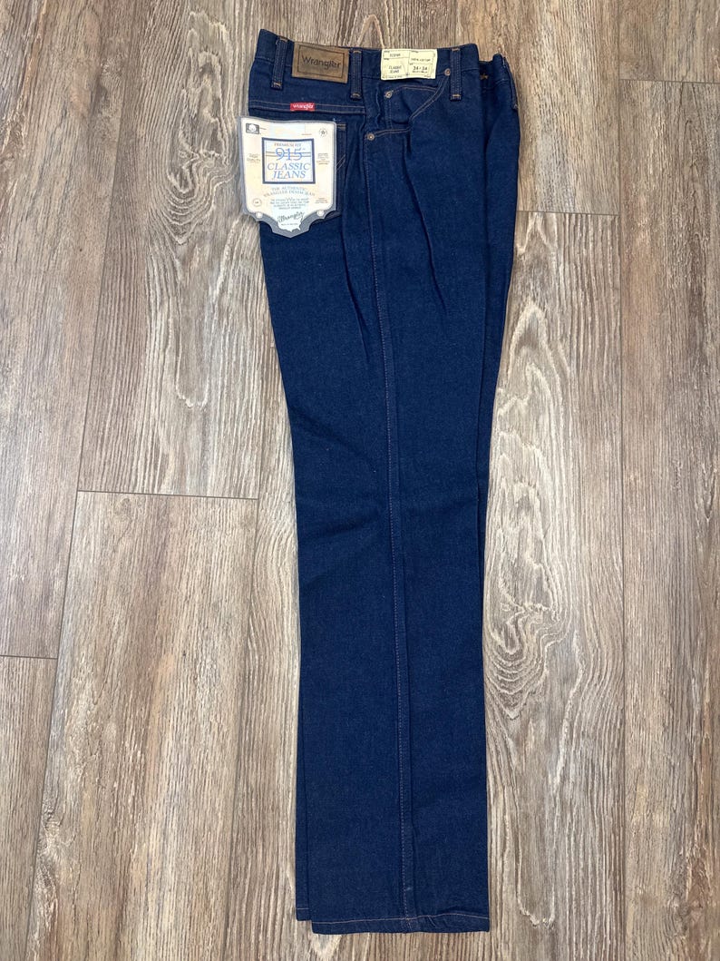 May include: Dark blue Wrangler 915 Classic Jeans, folded and displayed on a wooden surface. The jeans have a classic five-pocket design, a leather patch, and a tag that reads "915 Classic Jeans". The size tag reads 34x34.