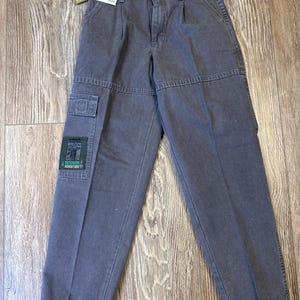 K&ouml;nnte beinhalten: Graue Wrangler Cargo-Hose mit mehreren Taschen und einem Aufn&auml;her mit der Aufschrift "Outdoor Adventure". Die Hose hat einen Knopf- und Rei&szlig;verschluss und wird auf einer Holzoberfl&auml;che pr&auml;sentiert. Ein Preisschild ist angebracht.
