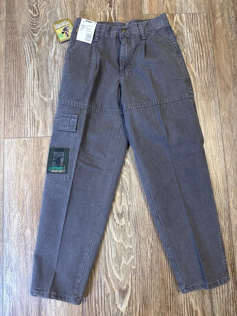 K&ouml;nnte beinhalten: Graue Wrangler Cargo-Hose mit mehreren Taschen und einem Aufn&auml;her mit der Aufschrift "Outdoor Adventure". Die Hose hat einen Knopf- und Rei&szlig;verschluss und wird auf einer Holzoberfl&auml;che pr&auml;sentiert. Ein Preisschild ist angebracht.