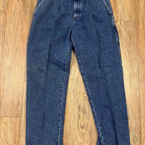 May include: Classic blue denim jeans with a button and zip fastening, front pleats, and pockets. The jeans have a straight leg and a vintage appearance. A brown tag is attached. The jeans are made of denim.