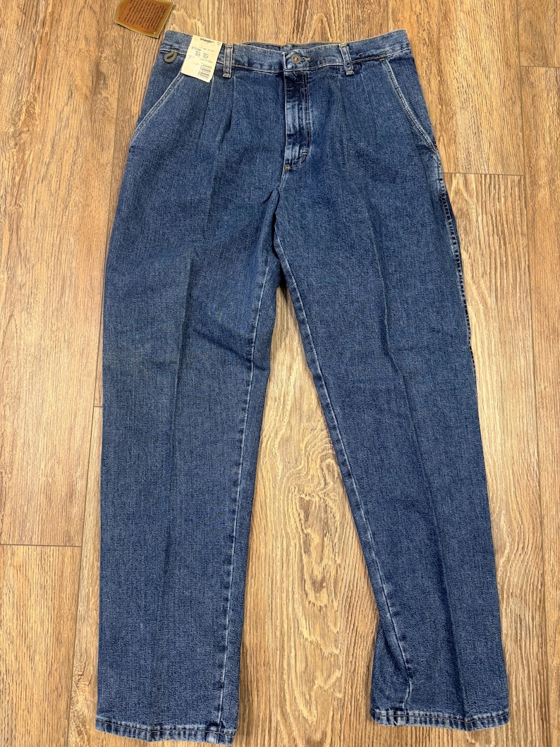 May include: Classic blue denim jeans with a button and zip fastening, front pleats, and pockets. The jeans have a straight leg and a vintage appearance. A brown tag is attached. The jeans are made of denim.