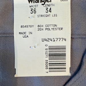 May include: A close-up of a Wrangler tag on gray denim pants. The tag indicates a waist size of 36 inches and a length of 34 inches. The pants are made of 80% cotton and 20% polyester. The tag also includes a barcode and the text "MADE IN USA."