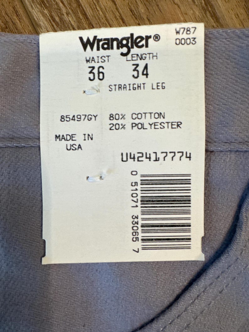 May include: A close-up of a Wrangler tag on gray denim pants. The tag indicates a waist size of 36 inches and a length of 34 inches. The pants are made of 80% cotton and 20% polyester. The tag also includes a barcode and the text "MADE IN USA."