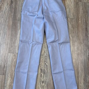 May include: Light blue Wrangler jeans, viewed from the back, with two back pockets and a small tag. The jeans are laid flat on a wooden surface. The tag reads "Wrangler".