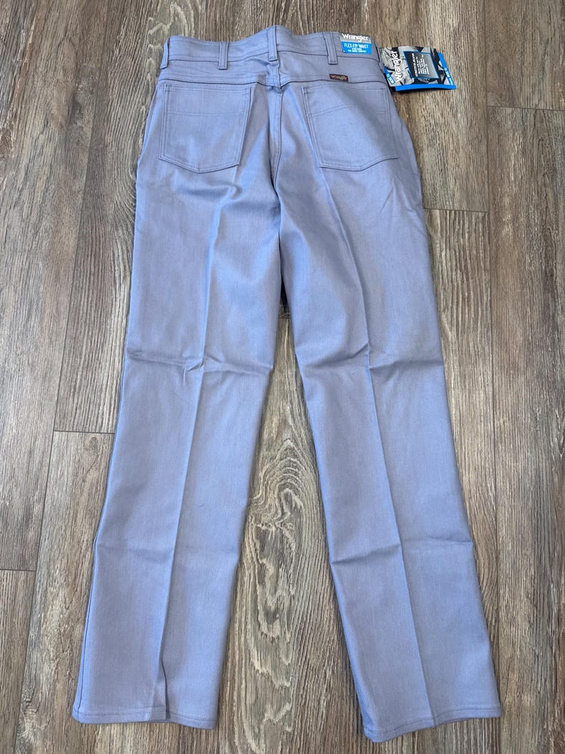 May include: Light blue Wrangler jeans, viewed from the back, with two back pockets and a small tag. The jeans are laid flat on a wooden surface. The tag reads "Wrangler".