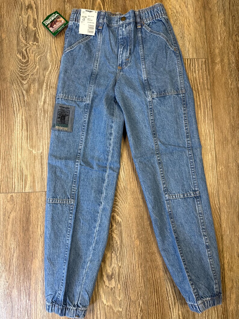K&ouml;nnte beinhalten: Hellblaue Jeanshose mit elastischem Bund und mehreren Taschen. Die Hose hat einen Aufn&auml;her mit der Aufschrift "Wrangler Outdoor Adventure". Die Hose hat eine lockere Passform und ist an den Kn&ouml;cheln ger&uuml;scht.