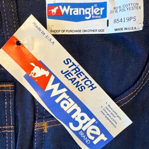 Peut inclure: Un jean en denim bleu avec une &eacute;tiquette blanche indiquant "Stretch Jeans" et "Wrangler Brand". L'&eacute;tiquette comprend &eacute;galement le texte "Move with you. Never bind. Give you a trim. Neat appearance". Le jean a une couture en fil brun et une &eacute;tiquette blanche avec le logo Wrangler et le texte "Full 1 Year Warranty" et "80% Cotton 20% Polyester". L'&eacute;tiquette comprend &eacute;galement la taille "34 x 32" et le texte "85419PS" et "Made in U.S.A."