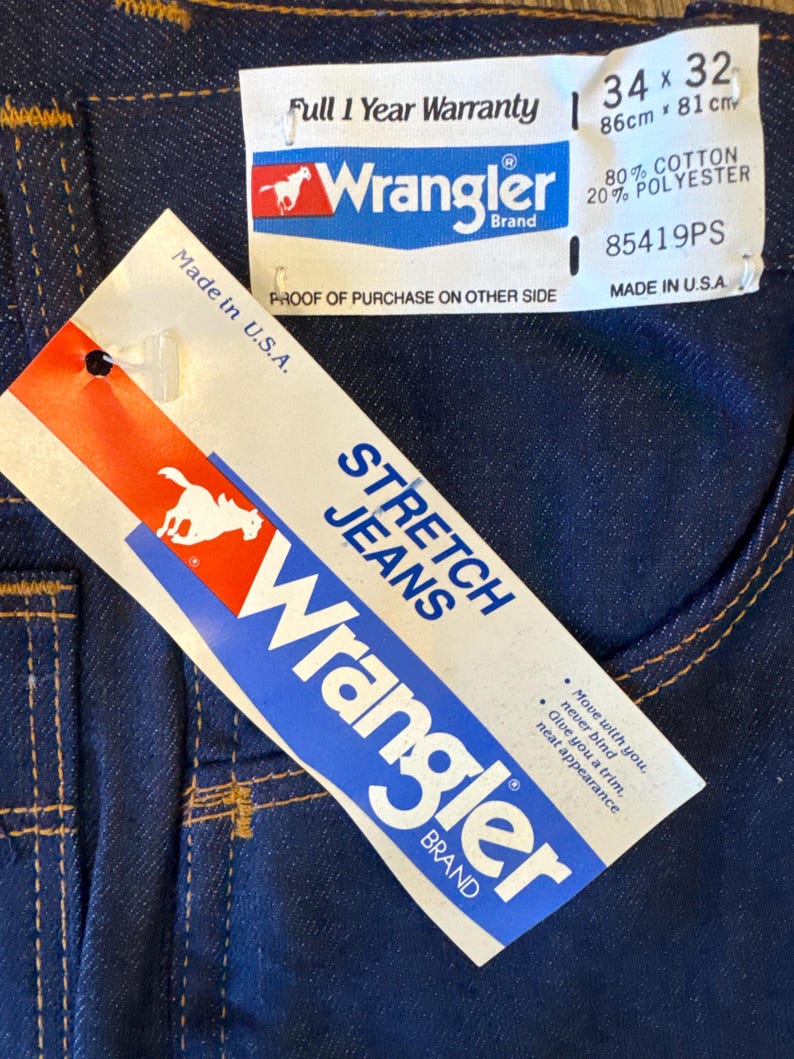 Peut inclure: Un jean en denim bleu avec une &eacute;tiquette blanche indiquant "Stretch Jeans" et "Wrangler Brand". L'&eacute;tiquette comprend &eacute;galement le texte "Move with you. Never bind. Give you a trim. Neat appearance". Le jean a une couture en fil brun et une &eacute;tiquette blanche avec le logo Wrangler et le texte "Full 1 Year Warranty" et "80% Cotton 20% Polyester". L'&eacute;tiquette comprend &eacute;galement la taille "34 x 32" et le texte "85419PS" et "Made in U.S.A."