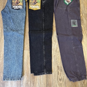 Może przedstawiać: Trzy pary dżins&oacute;w Wrangler Rugged Wear w r&oacute;żnych odcieniach. Dżinsy są niebieskie, czarne i szare. Każda para ma metkę Wrangler z grafiką osoby na polu. Dżinsy są prezentowane na drewnianej powierzchni.