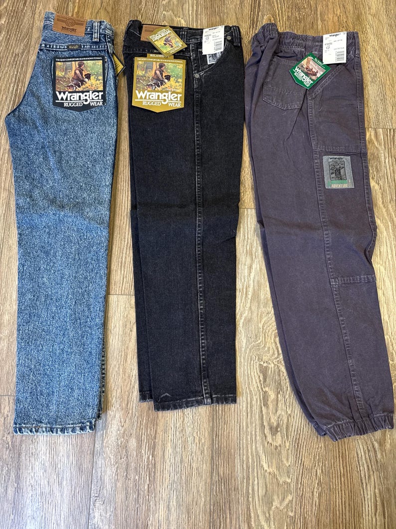 Może przedstawiać: Trzy pary dżins&oacute;w Wrangler Rugged Wear w r&oacute;żnych odcieniach. Dżinsy są niebieskie, czarne i szare. Każda para ma metkę Wrangler z grafiką osoby na polu. Dżinsy są prezentowane na drewnianej powierzchni.