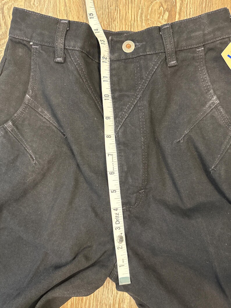 May include: Dark gray denim pants with a high waist and decorative stitching. A measuring tape shows a length of approximately 14 inches. The pants have a button closure and belt loops.