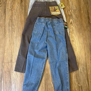 K&ouml;nnte beinhalten: Drei Jeanshosen von Wrangler in verschiedenen Waschungen: helles Acid-Wash, Dunkelbraun und Hellblau. Die Jeans haben elastische Bundweiten und Taschen. Die braune Hose hat einen Wrangler-Aufn&auml;her auf der Ges&auml;&szlig;tasche. Die Jeans werden auf einer Holzoberfl&auml;che pr&auml;sentiert.