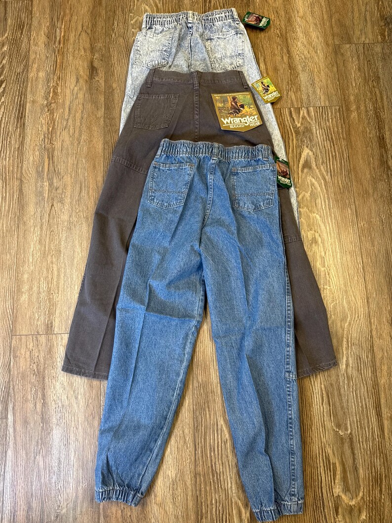 K&ouml;nnte beinhalten: Drei Jeanshosen von Wrangler in verschiedenen Waschungen: helles Acid-Wash, Dunkelbraun und Hellblau. Die Jeans haben elastische Bundweiten und Taschen. Die braune Hose hat einen Wrangler-Aufn&auml;her auf der Ges&auml;&szlig;tasche. Die Jeans werden auf einer Holzoberfl&auml;che pr&auml;sentiert.
