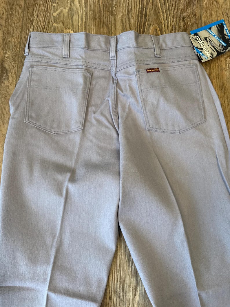 May include: Light blue Wrangler jeans with a classic five-pocket design. The jeans have a straight leg cut and a small red Wrangler logo patch on the back pocket. A blue and white tag is attached.