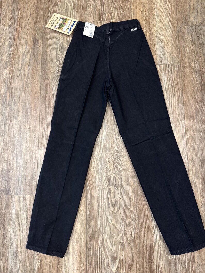 May include: Black Wrangler jeans with a straight leg design. The jeans have a classic five-pocket style and a high waist. A tag is attached to the jeans.