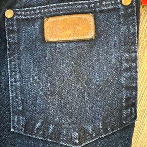 May include: Close-up of a dark wash denim Wrangler jeans pocket. The pocket features a brown leather patch with the Wrangler logo and "RIATA JEANS" text. Gold-colored rivets secure the pocket to the jeans. The Wrangler tag is visible at the top.