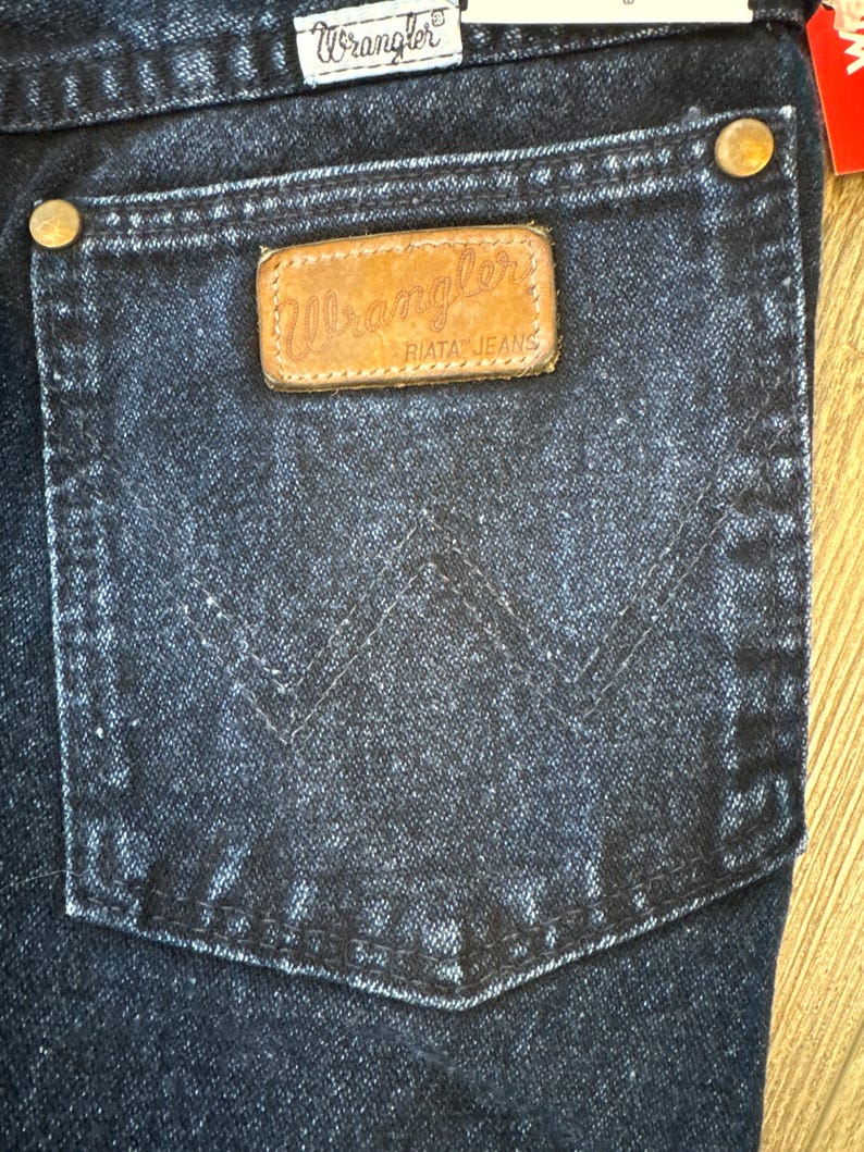 May include: Close-up of a dark wash denim Wrangler jeans pocket. The pocket features a brown leather patch with the Wrangler logo and "RIATA JEANS" text. Gold-colored rivets secure the pocket to the jeans. The Wrangler tag is visible at the top.