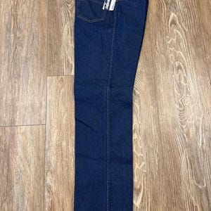 May include: Dark blue denim jeans with a straight leg cut. The jeans have a classic five-pocket design, a red Wrangler logo patch, and a hang tag. The jeans are displayed on a wooden surface.