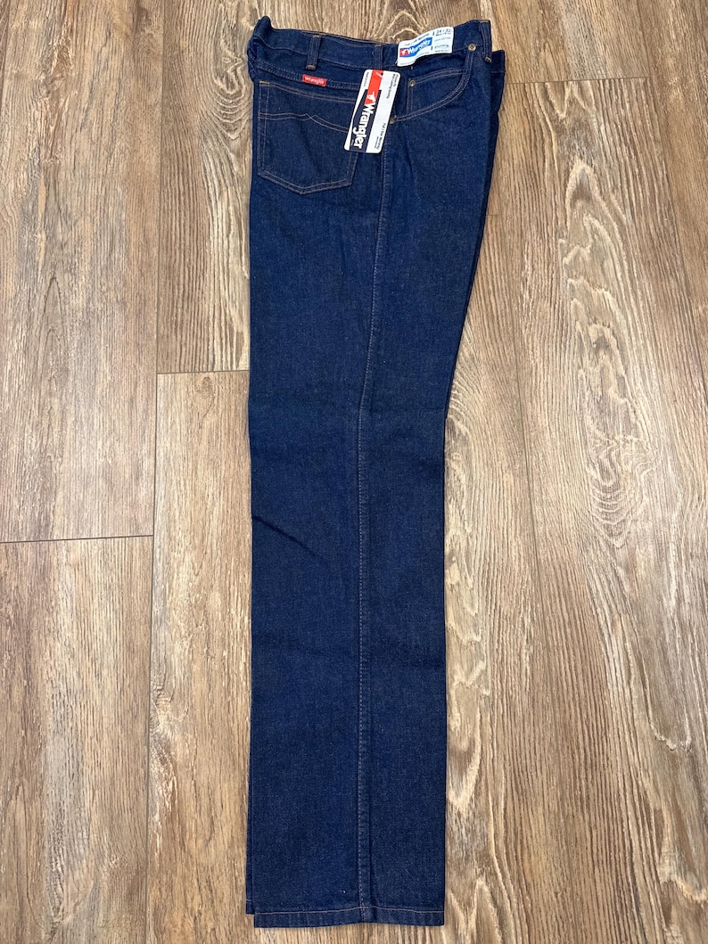 May include: Dark blue denim jeans with a straight leg cut. The jeans have a classic five-pocket design, a red Wrangler logo patch, and a hang tag. The jeans are displayed on a wooden surface.