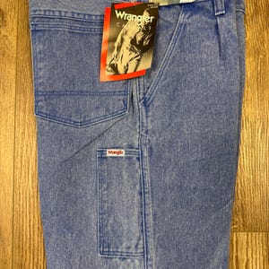 May include: Light blue Wrangler carpenter jeans with multiple pockets and a branded tag. The jeans are size 32x32 and made of denim. The tag features the Wrangler logo and a photo of a person.