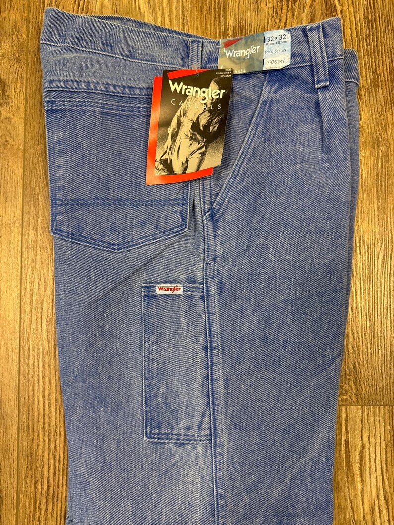 May include: Light blue Wrangler carpenter jeans with multiple pockets and a branded tag. The jeans are size 32x32 and made of denim. The tag features the Wrangler logo and a photo of a person.
