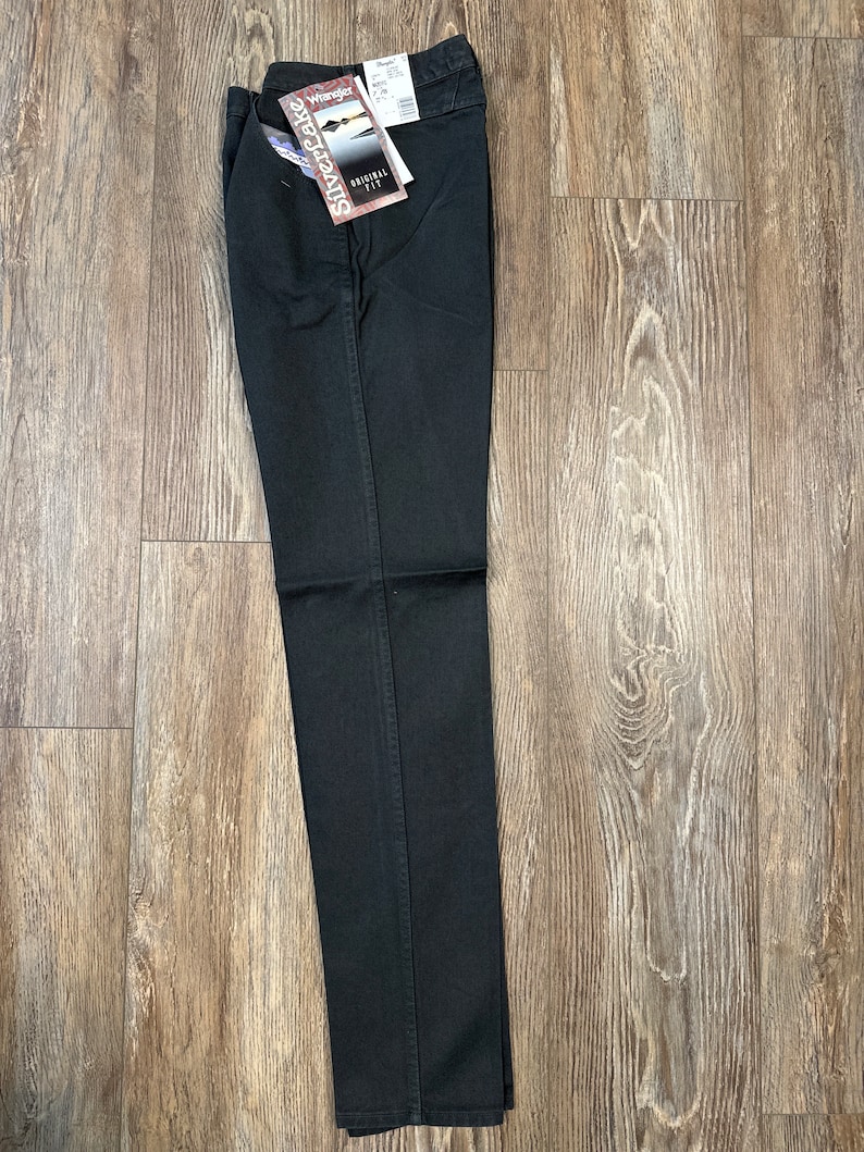 Op de afbeelding: Zwarte jeans met een Wrangler Silverlake Original Fit-label eraan bevestigd. De jeans is dubbelgevouwen en ligt plat op een houten vloer.