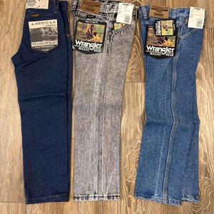 Puede incluir: Tres pares de pantalones vaqueros azules, uno azul oscuro, uno gris claro y uno azul claro. Los vaqueros son todos de la marca Wrangler y tienen diferentes etiquetas. Los vaqueros azul oscuro tienen una etiqueta que dice "American Legend Wrangler Blues". Los vaqueros gris claro tienen una etiqueta que dice "Wrangler Rugged Wear". Los vaqueros azul claro tienen una etiqueta que dice "Wrangler Rugged Wear".