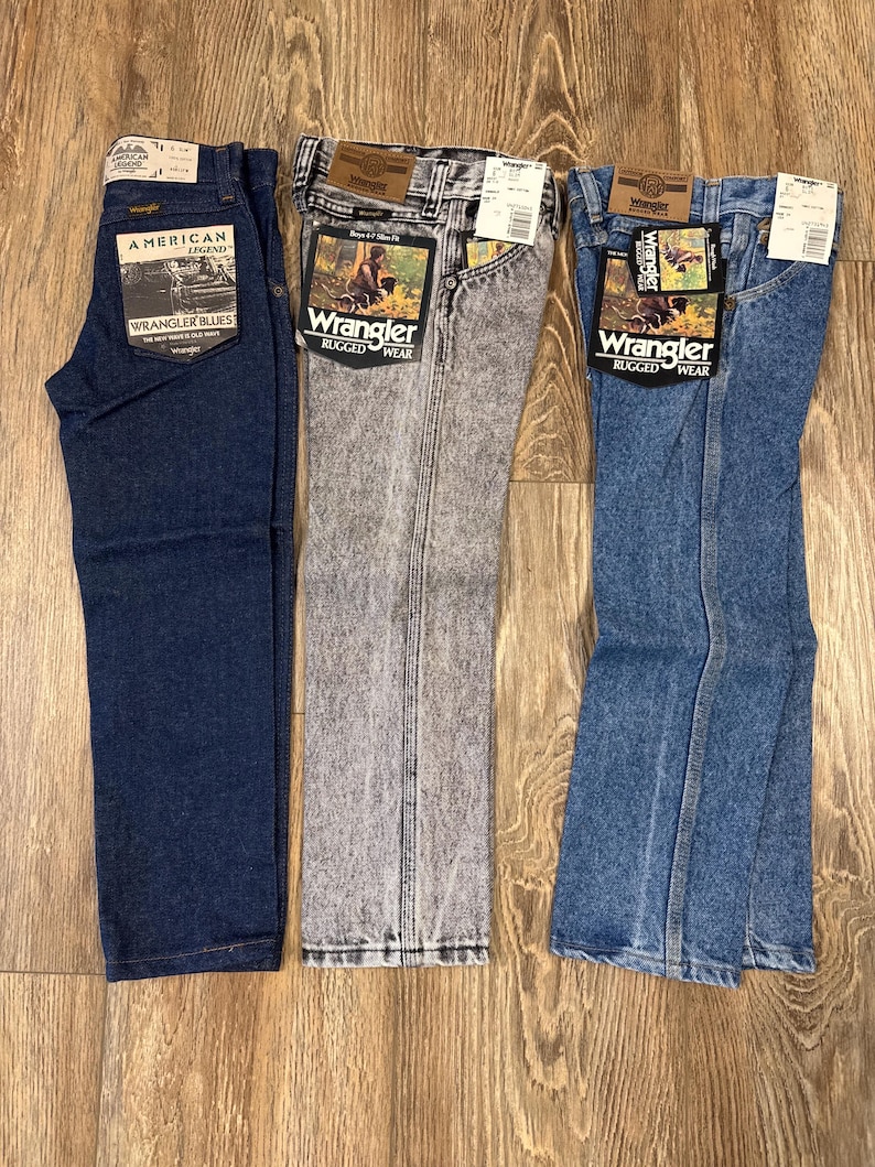 Puede incluir: Tres pares de pantalones vaqueros azules, uno azul oscuro, uno gris claro y uno azul claro. Los vaqueros son todos de la marca Wrangler y tienen diferentes etiquetas. Los vaqueros azul oscuro tienen una etiqueta que dice "American Legend Wrangler Blues". Los vaqueros gris claro tienen una etiqueta que dice "Wrangler Rugged Wear". Los vaqueros azul claro tienen una etiqueta que dice "Wrangler Rugged Wear".