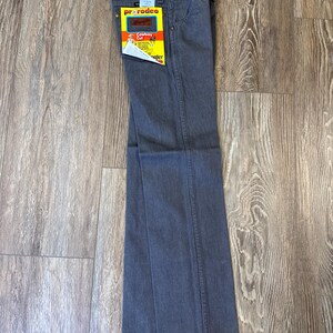 May include: Gray Wrangler Pro Rodeo Cowboy Cut jeans with a yellow and red tag. The jeans are folded and laid flat on a wooden surface. The tag features the Wrangler logo and product information.