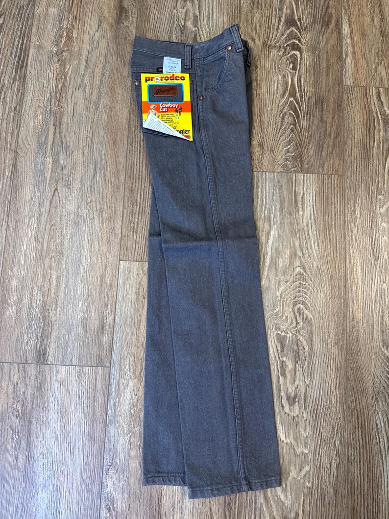 May include: Gray Wrangler Pro Rodeo Cowboy Cut jeans with a yellow and red tag. The jeans are folded and laid flat on a wooden surface. The tag features the Wrangler logo and product information.