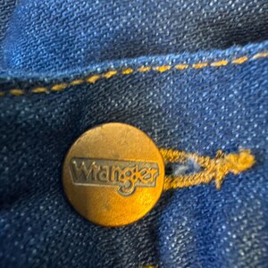 May include: Close-up of a blue denim jean with a gold Wrangler button.